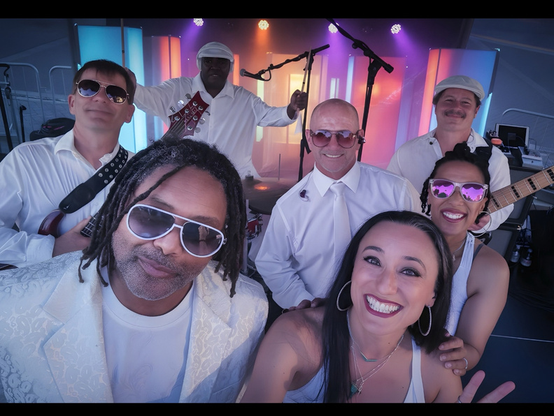 The Cheeseballs band members posing together for a group selfie in white outfits and sunglasses, with colorful stage lights behind them.